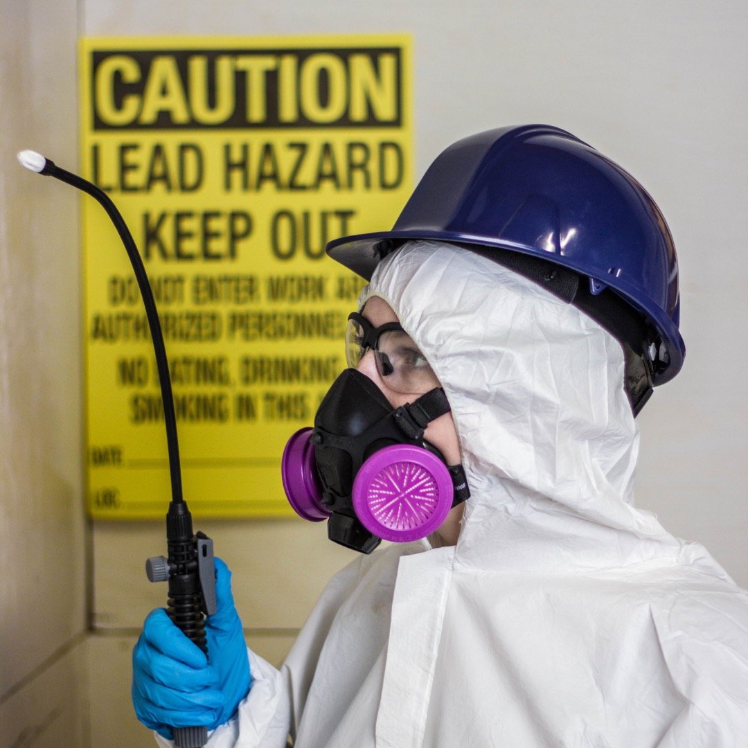 Someone in a hazard suit with a yellow “caution lead hazard” sign in the background.