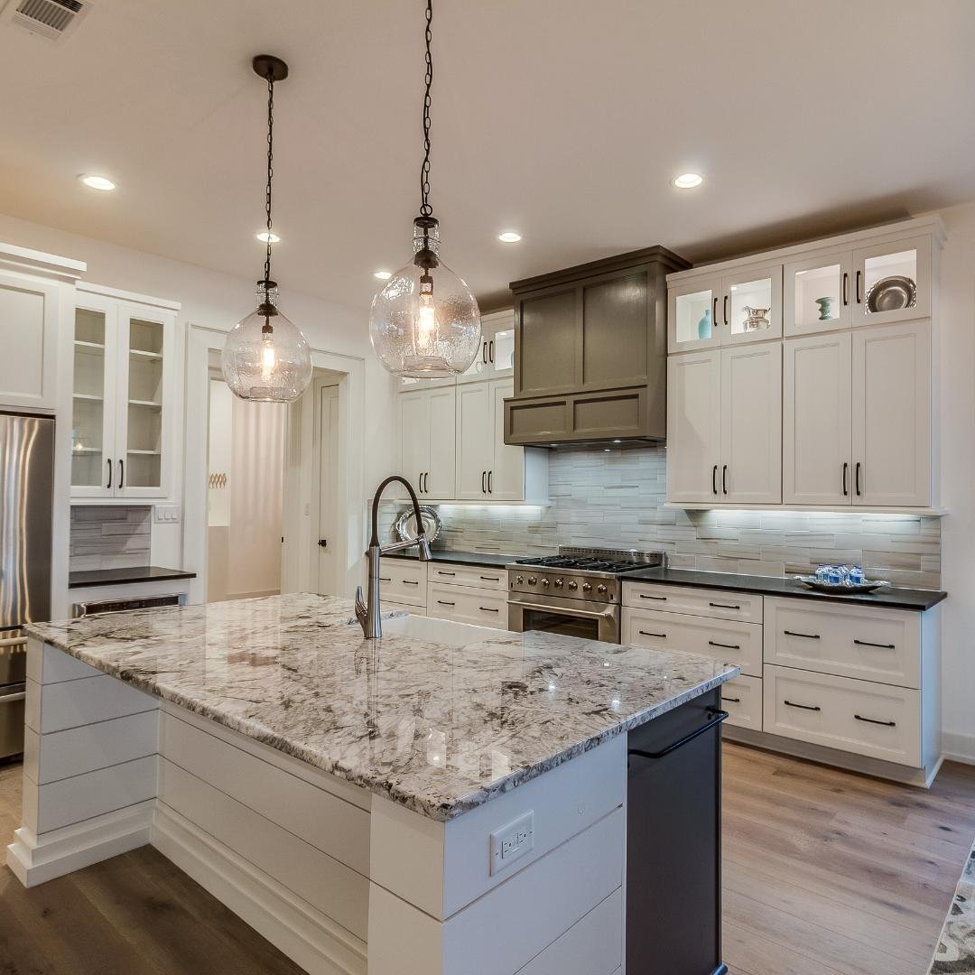 A close-up image of two pendant lights with clear glass globes being installed over a kitchen sink. The kitchen has white cabinets, granite countertops, and a stainless steel faucet.