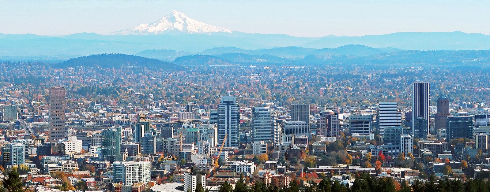 A panoramic view of the Portland, Oregon skyline at sunset, with snow-capped Mount Hood in the background.