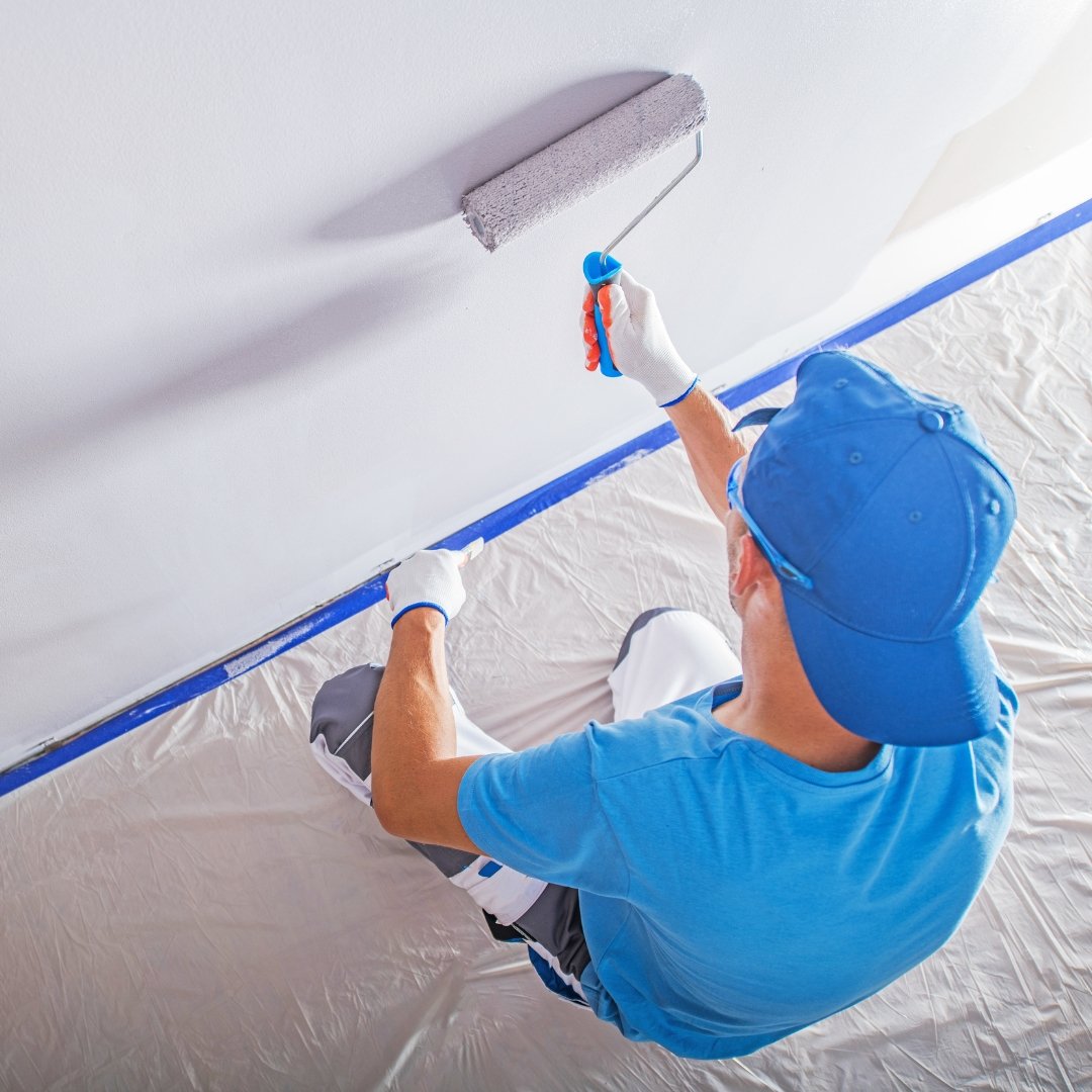 Gentleman painting the interior wall of a home. Wearing a blue shirt and cap.